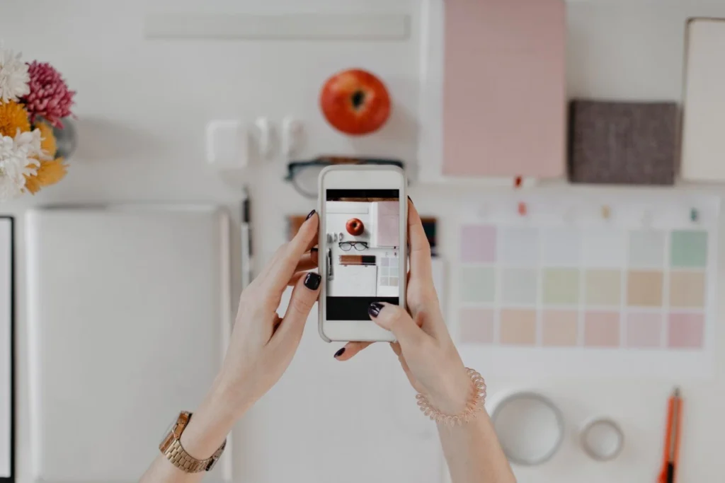 Pessoa segurando um celular tirando foto de uma mesa organizada, ideal para criar conteúdo para Instagram, com itens como maçã, flores e materiais de escritório.